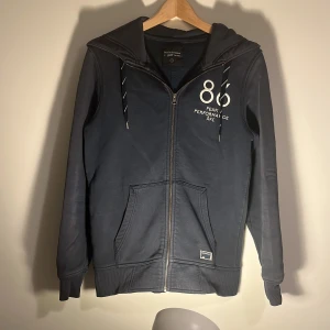 Mörkblå hoodie från Peak Performance - Säljer en snygg mörkblå hoodie från Peak Performance med dragkedja och tryck på bröstet. Den har en justerbar huva med snören och praktiska fickor framtill. S/M passar upp till 175 cm utan problem.