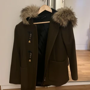 Brun kappa med fuskpäls från Zara Woman - Snygg brun kappa från Zara Woman med fuskpäls på huvan. Den har dekorativa spännen och dragkedja framtill. Perfekt för kyliga dagar med stil. 🧥✨Pris kan diskuteras :). 