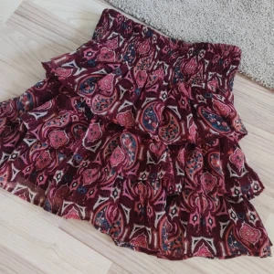 Mönstrad kortkjol med volanger - Säljer en färgglad kortkjol med ett livligt mönster i rött, rosa och blått. Kjolen har flera lager av volanger och en elastisk midja för bekväm passform. 💗