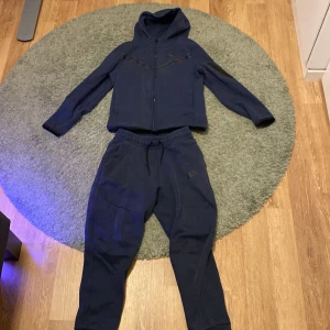 Nike tech - Säljer ett par mörkblå mjukisbyxor och hoodie från Nike med snörning i midjan och dragkedjefickor. Perfekta för en avslappnad stil. Byxorna har en broderad Nike-logga på sidan. Och hoodien har en ficka på armen, kan gå ner i pris