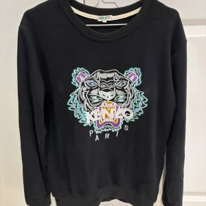 Svart sweatshirt från Kenzo - Snygg svart sweatshirt från Kenzo med ett coolt broderat tigerhuvud och texten 'Kenzo Paris' på framsidan. Tröjan har långa ärmar och en rund halsringning. Perfekt för en stilren och trendig look.