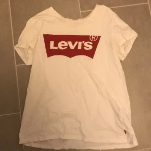 Vit t-shirt från Levi's - Säljer en klassisk vit t-shirt från Levi's med det ikoniska röda logotyptrycket på framsidan. T-shirten har en normal passform och är tillverkad i mjukt bomullsmaterial. Perfekt för en avslappnad stil. På bild 3 finns det ett lite hål men inget man tänker på🧜‍♀️🧜‍♀️
