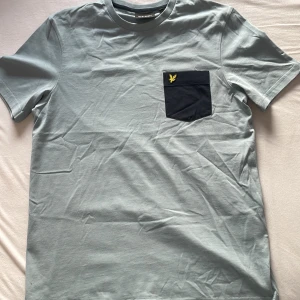 Grå t-shirt med ficka från Lyle & Scott - Snygg grå t-shirt från Lyle & Scott med en kontrasterande svart ficka på bröstet. Fickan har en broderad gul logotyp. Tillverkad i 100% bomull för en skön känsla. Perfekt för en avslappnad stil.