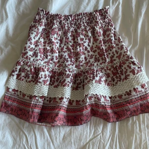 Blommig kjol från Vero Moda - Charmig blommig kjol från Vero Moda i vitt och rött. Kjolen har en volangdetalj och elastisk midja för en bekväm passform. Perfekt för varma dagar med sitt lätta och luftiga material.