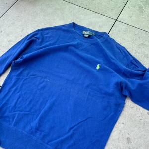 Blå tröja från Polo Ralph Lauren - Säljer en snygg blå tröja från Polo Ralph Lauren. Den har en klassisk design med långärmade ärmar och en liten grön logga på bröstet. Perfekt för både vardag och lite finare tillfällen. Tröjan är i bomull och känns riktigt skön att ha på sig.