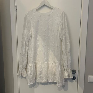 Viral studentklänning  - En vit klänning med långa ärmar och blommönster. Klänningen har en rund halsringning och en lös passform. Den har en volang vid nederkanten och är perfekt för festliga tillfällen. Använd 1 gång! Jag är 172 cm lång.