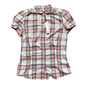 Rutig kortärmad skjorta från de.corp - Säljer en superfin rutig blus från de.corp i rosa, blå och vit. Blusen har korta ärmar och knappar hela vägen ner. Den har också snygga detaljer vid axlarna. Aldrig använd och lappen är fortfarande kvar. Nypris: 399 kr