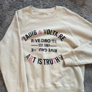 Beige sweatshirt från Zadig & Voltaire - Snygg beige sweatshirt från Zadig & Voltaire med tryck i svart, rosa och grönt. Texten 'Art is Truth' och en liten dödskalle ger en cool touch. Perfekt för en avslappnad stil.