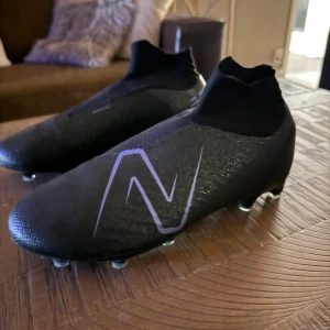 Svarta fotbollsskor från New Balance - Snygga svarta fotbollsskor från New Balance med en stilren design och diskreta lila detaljer. Skorna har en strumpliknande passform för extra komfort och stabilitet på planen. Perfekta för den som vill ha både stil och funktion i sitt spel. Ändas använda 1 gång. Vid frågor är det bara att skriva. MVH Felix