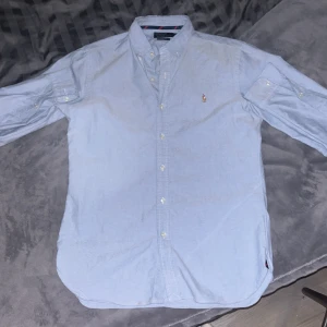 Ljusblå skjorta från Polo Ralph Lauren - Säljer en stilren ljusblå skjorta från Polo Ralph Lauren i slim fit. Skjortan har långa ärmar och knappar framtill samt en broderad logga på bröstet. Perfekt för en snygg och klassisk look.