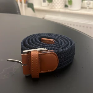 Blått flätat bälte 120 - Snyggt blått flätat bälte med bruna läderdetaljer och en silverfärgad spänne. Perfekt för att ge en stilren touch till din outfit. Bältet är 120 cm långt och passar de flesta storlekar.