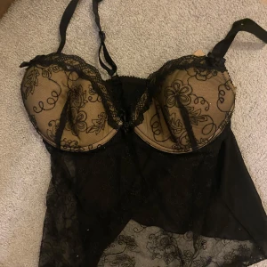 Svart spetskorsett med blommönster - Elegant svart korsett med blommönster och spetsdetaljer. Den har justerbara axelband och en transparent design. Perfekt som fest topp