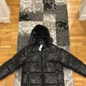 Svart dunjacka från Polo Ralph Lauren - Snygg svart dunjacka från Polo Ralph Lauren med ikonisk röd logga på bröstet. Jackan har en praktisk dragkedja och en huva för extra skydd mot väder. Perfekt för kyliga dagar. Pris kan diskuteras 