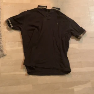 Svart piké från Atlant - Säljer en svart piké  från Atlant med en liten broderad logga på bröstet. Piken har en klassisk krage med knappar och vita ränder på ärmsluten. Perfekt för ett kalas. Storlek L passar M