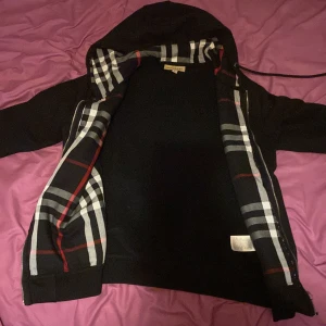 Svart hoodie från Burberry - Säljer en snygg svart hoodie från Burberry med klassiskt rutigt mönster på insidan av huvan. Jackan har en dragkedja framtill och en diskret broderad logga på bröstet. Perfekt för en stilren look!