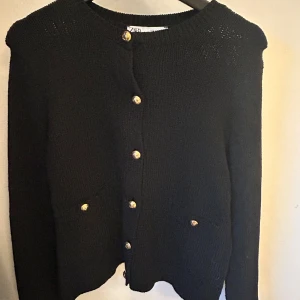 Svart kofta med guldknappar - Elegant svart kofta från Zara med dekorativa guldknappar framtill. Den har långa ärmar och två fickor fram. Perfekt för en stilren look.