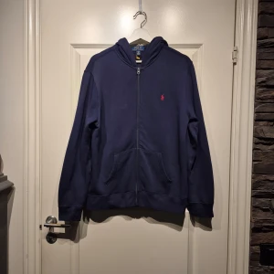 Mörkblå hoodie från Ralph Lauren - Säljer en mörkblå hoodie från Ralph Lauren med dragkedja. Broderad röd logga på bröstet. Mycket fint skick. Ni är alltid välkomna att höra av er vid eventuella frågor eller funderingar. 