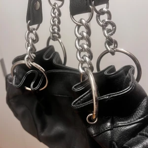 Svart axelväska från River Island - Snygg svart axelväska från River Island med kedjedetaljer och rundade handtag. Väskan har en stilren design och är perfekt för att bära dina dagliga nödvändigheter. Den är tillverkad i skinnliknande material och har en praktisk storlek.