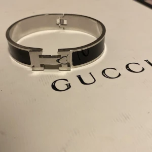 H armband - Elegant armband  i svart och silver. Armbandet har en stilren design med en metallisk finish och ett ikoniskt spänne. Perfekt för att ge en lyxig touch till din stil.