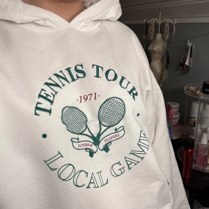 Vit hoodie med tennistryck - Säljer en vit hoodie med tryck 'Tennis Tour Local Game' och två tennisracketar. Perfekt för en sportig look eller avslappnad stil. Hoodien har en klassisk passform med långa ärmar och en bekväm huva.