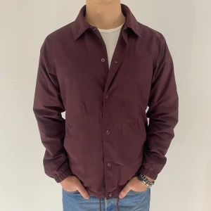 Vinröd jacka från Dickies - Jackan har en klassisk krage och stängs med knappar. Den har långa ärmar med elastiska muddar och dragsko i nederkanten för justerbar passform. Storlek medium. Modellen är 180cm och väger 75kg. 