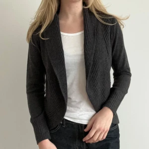 Mörkgrå kofta - Säljer en stilren mörkgrå kofta med ribbade detaljer. Koftan har långa ärmar och en öppen design framtill, perfekt för lager-på-lager. Passar både till vardags och mer uppklädda tillfällen.