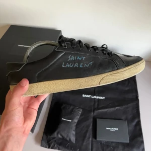 Saint Laurent skor - Snygga svarta sneakers från Saint Laurent med handskriven logga på sidan. Skorna har en klassisk design med snörning och en ljus sula som ger en cool kontrast. Perfekta för en stilren look.