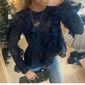 marinblå blus med volanger - Snygg marinblå blus med volanger och långärmad design. Perfekt för en stilren look med en touch av elegans. Blusen har ett subtilt mönster och är tillverkad i ett lätt material.