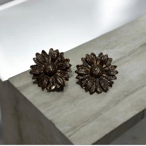 Blommiga örhängen i brons (clips) - Snygga örhängen i form av blommor med en bronsfärgad finish. De har en detaljrik design med kronblad som ger en elegant och unik look. Perfekta för att ge en extra touch till din outfit.