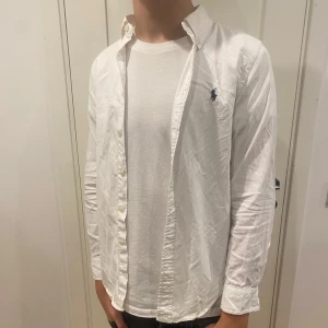 Vit skjorta från Ralph Lauren - Klassisk vit skjorta från Ralph Lauren med broderad logga på bröstet. Skjortan har långa ärmar och knappar framtill. Perfekt för en stilren look. Storlek 12 barn sitter som xs 