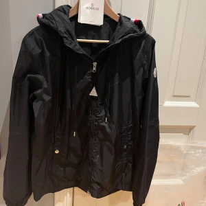 Svart vindjacka från Moncler - Säljer en stilren svart vindjacka från Moncler med huva och dragkedja. Jackan har Monclers logotyp på ärmen och röd-vit randdetalj på huvan. Perfekt för blåsiga dagar. Är bara använd 5 gånger är som ny och alla svanar funkar som ni ser på bilderna bara skicka vid minsta fråga stl 2 som motsvarar M priset kan diskuteras vid snabb och smidig affär 