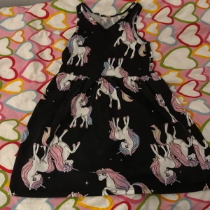 Svart klänning med enhörningsmönster - Charmig svart klänning med ett färgglatt enhörningsmönster. Klänningen har en ärmlös design och en rund halsringning. Perfekt för den som älskar magiska motiv och vill sticka ut med en unik stil.