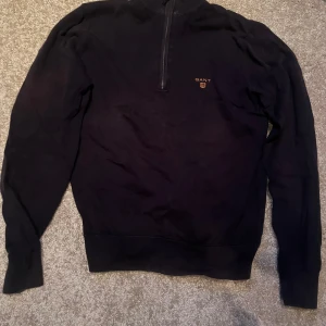 Svart tröja med dragkedja från Gant - Säljer en stilren svart tröja från Gant med halv dragkedja och broderad logga på bröstet. Perfekt för en avslappnad men snygg look. Tröjan har långa ärmar och är tillverkad i ett mjukt material.