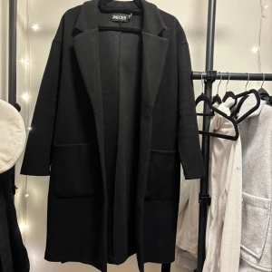 Svart kappa från Pieces - Snygg svart kappa från Pieces med knytband i midjan. Den har långa ärmar och två stora fickor framtill. Perfekt för en stilren look. nypris 549. anvånd fåtal ggr