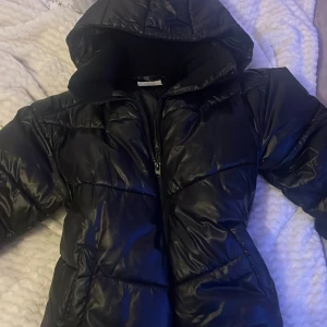 Svart pufferjacka från Lindex - Säljer en svart pufferjacka från Lindex för barn, storlek EUR 158. Jackan är vattenavvisande och har en huva samt dragkedja framtill. Perfekt för kyliga dagar! Har använt den några gånger men den var för stor. 