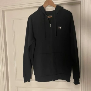 Svart hoodie från Under Armour - Säljer en svart hoodie från Under Armour med dragkedja och huva. Den har en liten logotyp på bröstet och är tillverkad i ett mjukt material. Jag säljer den för att den är för lite. Skriv tlll mig för ytterligare frågor.