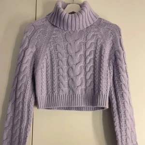 Lila stickad polotröja - Säljer en snygg lila stickad polotröja med flätmönster. Tröjan är croppad och har långa ärmar, perfekt för kyligare dagar. Den höga kragen ger extra värme och stil. Passar utmärkt till både jeans och kjol.