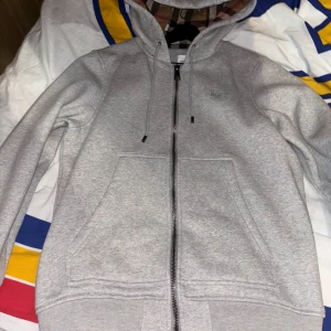 Grå huvtröja från Burberry - Säljer en stilren grå huvtröja från Burberry med dragkedja och klassiskt rutigt mönster i huvan. Tröjan har långa ärmar och en broderad logotyp på bröstet. Perfekt för en avslappnad och trendig look. Storlek L och är använd 1 gång. Priset kan alltid diskuteras