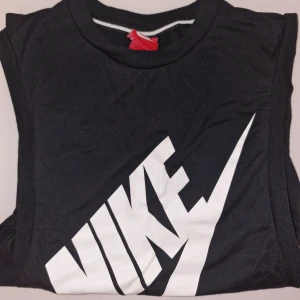 Svart träningslinne från Nike - Säljer ett svart träningslinne från Nike med stort vitt logotryck på framsidan. Perfekt för träning eller en avslappnad stil. Garderobsrensning så kika gärna på övriga auktioner.