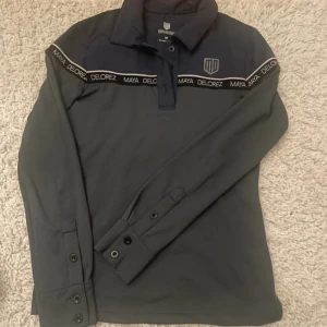 Mörkblå pikétröja från Maya Delorez - Snygg mörkblå pikétröja från Maya Delorez med långa ärmar och knappar framtill. Tröjan har ett stilrent band med logotypen runt bröstet och en diskret logga på bröstet. Perfekt för en sportig look.