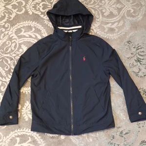 Mörkblå jacka från Polo Ralph Lauren - En mörkblå polo ralph lauren jacka med en tunn luva perfekt till våren. Jackan är använd ett par gånger, inga defekter på Jackan. Den är size m barnstorlek 11-12 år, passar även de som är 13 år. Priset går diskutera!