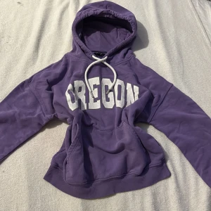 Lila hoodie från Don't Call Me Jennyfer - Säljer en snygg lila hoodie med texten 'OREGON' i vitt på framsidan. Den har en klassisk känguruficka och justerbar huva med vita snören. Perfekt för en avslappnad stil!