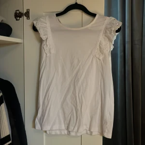 Vit ärmlös topp med spetsdetaljer - Säljer en söt vit ärmlös topp med spetsdetaljer på axlarna. Perfekt för varma dagar eller som ett lager under en kofta. Toppen har en rund halsringning och är gjord i ett mjukt material.