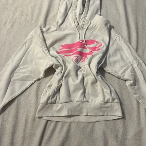 Vit hoodie från Weekday med rosa tryck - Säljer en vit hoodie från Weekday i storlek XS. Den har ett coolt rosa tryck på framsidan med japanska tecken och en figur. Hoodien har en klassisk design med dragsko i huvan och ribbade muddar.