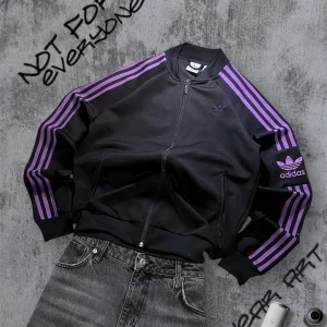 Svart och lila jacka från Adidas - Snygg svart tröja från Adidas med lila ränder längs ärmarna och det klassiska Adidas-loggan broderad på bröstet. Tröjan har en dragkedja framtill. Perfekt för en sportig look! Passar som en vanlig S