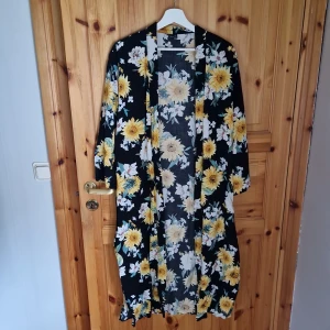 Blommig lång kimono - Säljer en superfin lång kimono med ett blommigt mönster i gult och vitt på en svart bakgrund. Den har trekvartsärmar och ett knytband i midjan. Perfekt för sommarens fester eller som en snygg detalj över en enkel outfit!