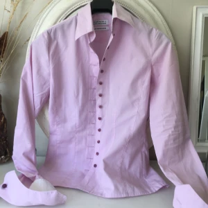 Linnéa Braun. skjorta från The Shirt Factory - Säljer en stilren ljusrosa skjorta från The Shirt Factory, designad av Linnéa Braun. Skjortan är i 100% bomull och är lätt att stryka. Den har långa ärmar och knappar framtill. Perfekt för både vardag och fest!