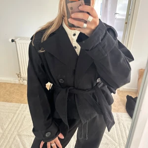 Kort trenchcoat Gina tricot  - Säljer denna trendiga svarta korta trenchcoat från Gina tricot. Den har knappar och ett snöre som går att knyta runt midjan, men funkar även att ha utan🥰  Köpt i våras! Strl XS💕
