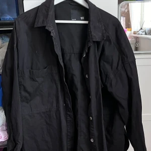 Svart overshirt - Säljer en svart overshirt. Den har en avslappnad passform och är perfekt för lager-på-lager-stil. Skjortan har knappar framtill och är långärmad. Perfekt för både vår och höst!
