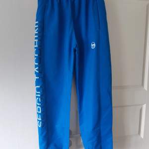 Säljer ett par snygga blå träningsbyxor från Sergio Tacchini. De har en elastisk midja och är dekorerade med märkesnamnet längs benet. Perfekta för träning eller en avslappnad dag. Byxorna är endast använda 2 gånger och därmed i fint skick och redo för nya äventyr! XL i storlek men mer som en medium (M) i verkligheten.